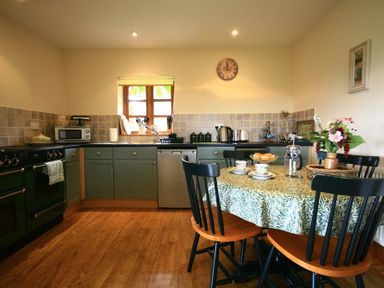 A kitchen with green cabinets a wooden floor a round table with a patterned tablecloth and black chairs at The Piggery Lydney