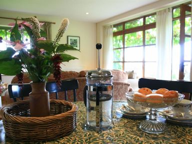 A dining table with a flower vase in a wicker basket a French press coffee maker and a glass pedestal bowl with muffins in a living room at The Piggery in Lydney
