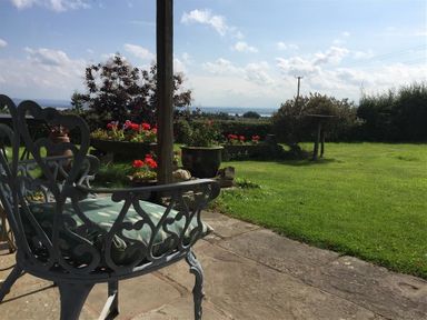 A garden with metal chairs on a stone patio and flower beds on a lawn at The Piggery in Lydney