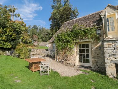 An exterior view of a stone cottage with a small garden table and chairs on grass at Mayfly Cottage Coln St Aldwyns near Bibury