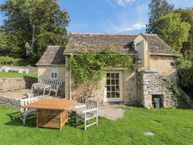 An outdoor seating area with wooden chairs and a table in front of a stone cottage with a tiled roof and greenery at Mayfly Cottage Coln St Aldwyns near Bibury
