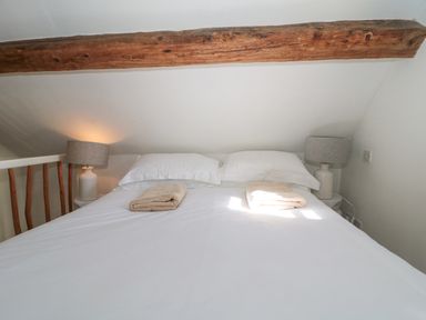 A bed with white sheets pillows and two folded towels under a wooden beam with lamps on either side at Mayfly Cottage in Coln St Aldwyns near Bibury