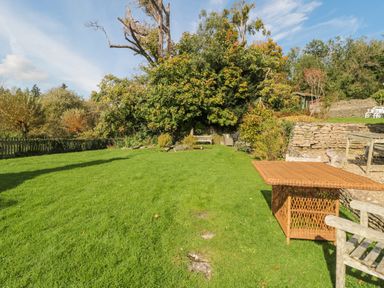 A garden with green lawn and trees with wooden furniture and wicker table at Mayfly Cottage Coln St Aldwyns near Bibury