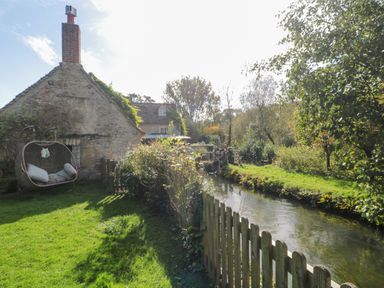 A stone cottage with a hanging chair in the garden next to a stream and wooden fence at Mayfly Cottage in Coln St Aldwyns near Bibury