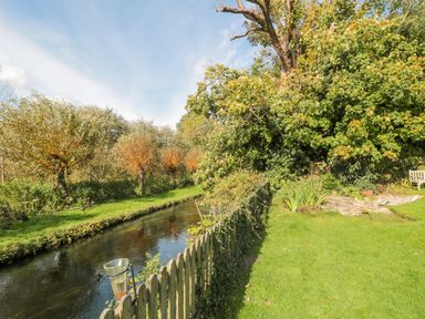 A grassy garden with a wooden fence beside a narrow river and trees at Mayfly Cottage in Coln St Aldwyns near Bibury