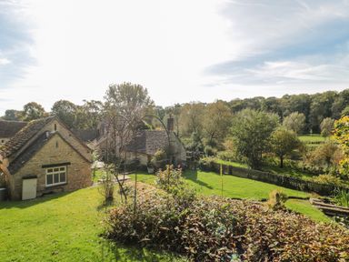 A garden with shrubs and trees near stone cottages with pitched roofs at Mayfly Cottage in Coln St Aldwyns near Bibury