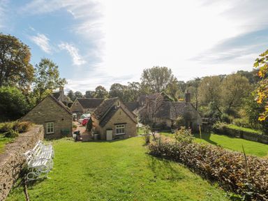 A garden with green grass white benches stone cottages and trees at Mayfly Cottage Coln St Aldwyns near Bibury