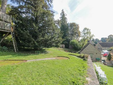 A grassy garden with trees a wooden treehouse and stone buildings at Mayfly Cottage in Coln St Aldwyns near Bibury