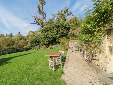 An outdoor garden area with wooden tables and chairs on grass and gravel path surrounded by trees and bushes at Mayfly Cottage in Coln St Aldwyns near Bibury