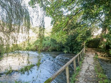 A narrow stone path with a wooden railing alongside a flowing river surrounded by trees and greenery at Mayfly Cottage in Coln St Aldwyns near Bibury