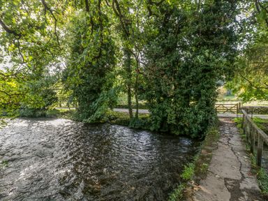 A river flowing next to a stone path and leafy trees at Mayfly Cottage in Coln St Aldwyns near Bibury