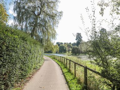 A pathway with a hedge on one side and a wooden fence bordering a grassy area with trees and a pond at Mayfly Cottage in Coln St Aldwyns near Bibury