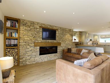A living room with two sofas a stone wall with a mounted TV and fireplace and a kitchen area at Old Groom's Cottage in Minster Lovell