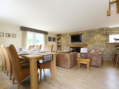 A dining area with a table set for six and a living room with sofas and a stone wall with a TV at Old Groom's Cottage in Minster Lovell