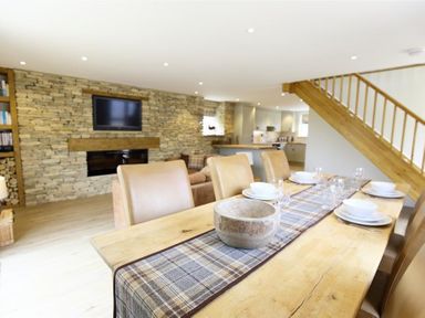A dining area with a wooden table set with plates and glasses next to a living room with a stone wall fireplace and a TV at Old Groom's Cottage in Minster Lovell