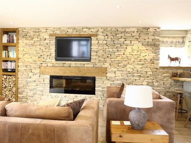 A living room with brown sofas a stone wall with a mounted television and fireplace a wooden side table with a lamp and built in bookcase at Old Groom's Cottage in Minster Lovell