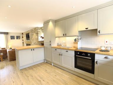 A kitchen with cabinets countertop and oven adjacent to a living area with stone wall and television at Old Groom's Cottage in Minster Lovell