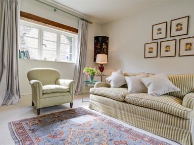 A living room with an armchair, a striped sofa with cushions, a rug, a side table with flowers and a lamp, and framed pictures on the wall at The Old Dairy in Shilton