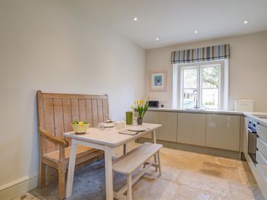 A kitchen with a wooden bench and white table with flowers near a window at The Old Dairy in Shilton