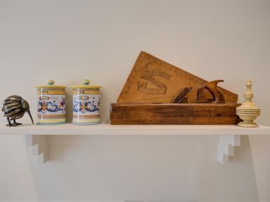 A white shelf with two painted jars a metal bird sculpture a wooden tool with a leather ruler and a cream decorative object at The Old Dairy in Shilton