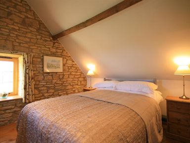 A bedroom with a double bed bedside tables with lamps a window with curtains and a stone wall at Calcot Peak Barn Bibury near Northleach