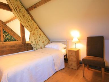 A bedroom with a single bed beside a wooden nightstand with a lamp and a brown chair at Calcot Peak Barn in Bibury near Northleach