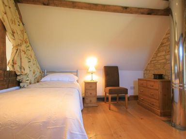 A bedroom with a single bed a wooden nightstand with a lamp a brown chair and a wooden dresser at Calcot Peak Barn Bibury near Northleach