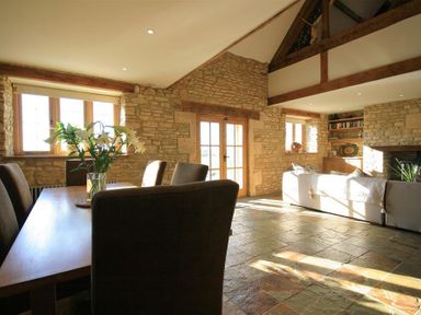 A living and dining area with a wooden table and chairs a sofa and stone walls at Calcot Peak Barn Bibury near Northleach