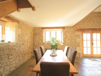 A dining room with a wooden table and six chairs and a vase with flowers on the table at Calcot Peak Barn in Bibury near Northleach