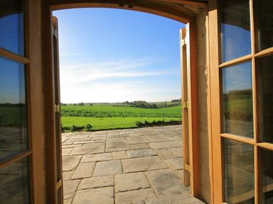 A stone patio outside open wooden double doors with a view of green fields and blue sky at Calcot Peak Barn in Bibury near Northleach
