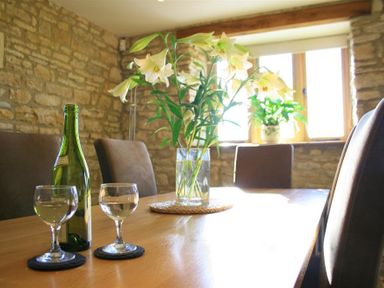 A dining table with a vase of flowers wine bottle and two glasses in a room with stone walls at Calcot Peak Barn Bibury near Northleach