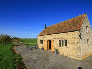 A stone house with wooden doors and windows on a paved patio in a rural area at Calcot Peak Barn in Bibury near Northleach
