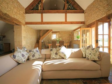 A living room with a beige sectional sofa and floral cushions next to wooden stairs and a dining table with chairs at Calcot Peak Barn Bibury near Northleach