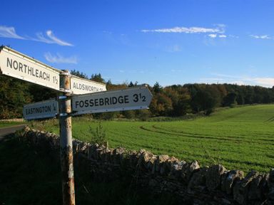 A rural road signpost at a crossroads with directions to Northleach Aldsworth Eastington and Fossebridge near green fields and trees at Calcot Peak Barn Bibury near Northleach
