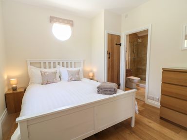 A bedroom with a white bed with floral pillows bedside tables lamps wooden chest of drawers and an en suite bathroom at Nutchat Barn in Burford