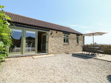 A stone exterior cottage with green doors and windows and a patio table with chairs and umbrella at Nutchat Barn in Burford