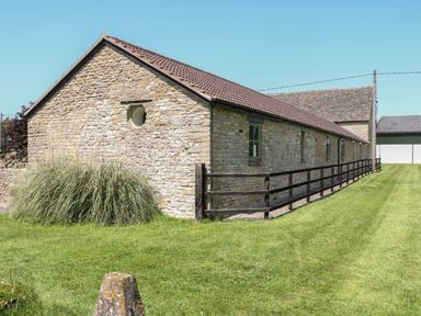A stone barn with a tiled roof and wooden fence on a lawn at Nutchat Barn in Burford