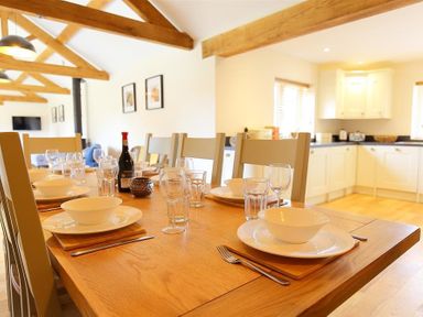 A dining table set with plates bowls glasses and cutlery in a room with exposed wooden beams and a kitchen in the background at Court Hayes Farm Barns Bourton-On-The-Water