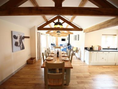 A dining room with a wooden table and chairs set with plates and glasses in an open space with exposed wooden beams and a kitchen area at Court Hayes Farm Barns in Bourton-On-The-Water