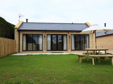 A single-story stone building with large black-framed windows and central doors facing a grassy yard with a wooden picnic table and umbrella at Court Hayes Farm Barns in Bourton-On-The-Water