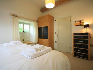 A bedroom with a bed with white linens towels on the bed a window with curtains a wooden wardrobe a door a chest of drawers with a lamp and artwork on the wall at Stepwell in Moreton-In-Marsh