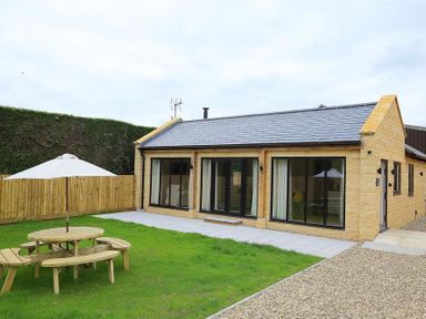 A single-story stone building with large windows a picnic table with an umbrella on a grassy yard at Park Bothy Wyck Rissington