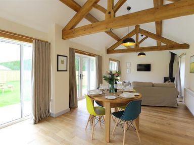 A dining area with a round wooden table and colorful chairs next to a living area with a sofa and a wood stove at Park Bothy in Wyck Rissington