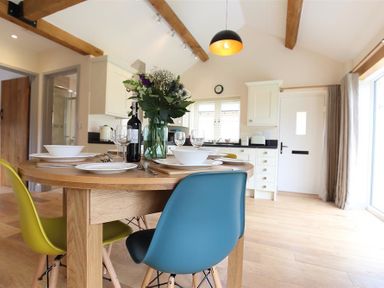 A dining area with a round wooden table set with plates and glasses surrounded by colorful chairs in a kitchen with exposed wooden beams at Park Bothy in Wyck Rissington