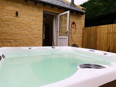 An outdoor hot tub next to a stone building with an open door and a wooden fence in the background at Park Bothy in Wyck Rissington