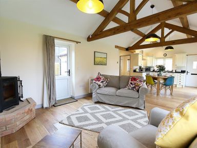 A living room with exposed wooden beams a gray sofa with floral cushions a dining table with colorful chairs and a kitchen in the background at Park Bothy in Wyck Rissington