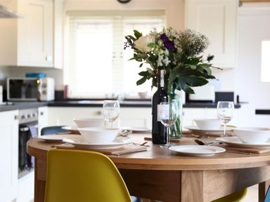 A kitchen with a wooden dining table set with white plates bowls wine glasses a bottle of wine and a vase of flowers at Park Bothy Wyck Rissington