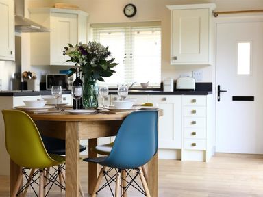 A kitchen with a wooden dining table set for four with colorful chairs and a flower vase at Park Bothy Wyck Rissington