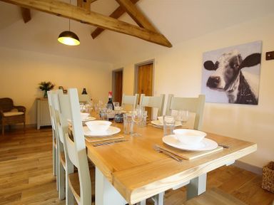 A dining room with a wooden table set with plates and glasses and a cow painting on the wall at Park Stables Wyck Beacon near Bourton-on-the-Water