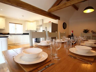 A dining table set with plates glasses and cutlery in a kitchen with wooden beams and white cabinets at Park Stables in Wyck Beacon near Bourton-on-the-Water